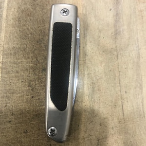 May include: A silver folding knife with a black rubber grip. The knife has a stainless steel blade and is secured with two screws. The handle is rectangular with rounded edges.
