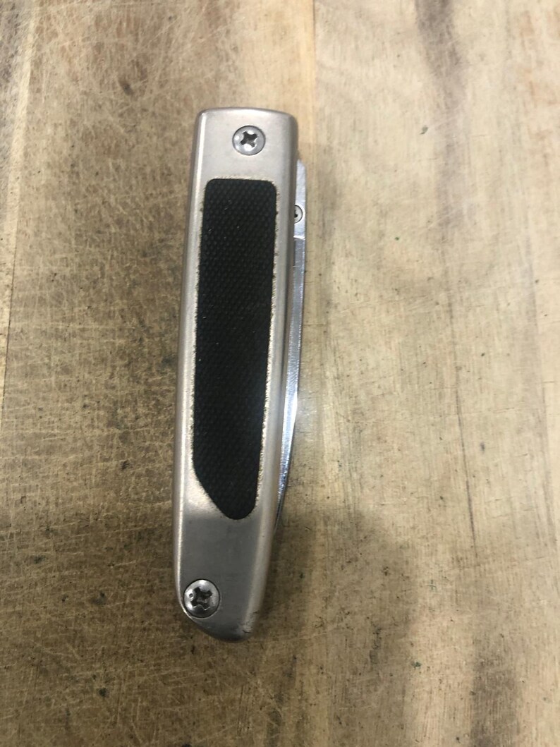 May include: A silver folding knife with a black rubber grip. The knife has a stainless steel blade and is secured with two screws. The handle is rectangular with rounded edges.
