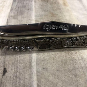 May include: A pocket knife with a stainless steel blade and a decorative handle. The handle features an ornate design with a fish and floral pattern. The knife also includes a corkscrew. The text "Fighter Plus" is engraved on the blade.