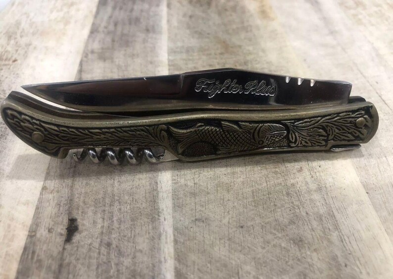 May include: A pocket knife with a stainless steel blade and a decorative handle. The handle features an ornate design with a fish and floral pattern. The knife also includes a corkscrew. The text "Fighter Plus" is engraved on the blade.
