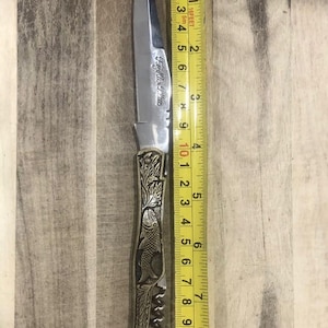 May include: A stainless steel pocket knife with a decorative handle featuring fish and floral engravings. The blade is approximately 4 inches long. The knife has the text "Fey Mles Plus" engraved on the blade.