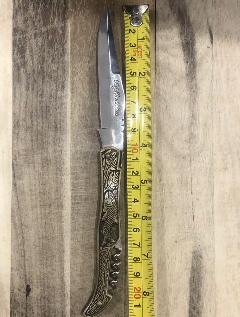 May include: A stainless steel pocket knife with a decorative handle featuring fish and floral engravings. The blade is approximately 4 inches long. The knife has the text "Fey Mles Plus" engraved on the blade.