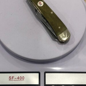 Op de afbeelding: Een gesloten zakmes met een groene handgreep en zilveren accenten, liggend op een weegschaal. Het mes heeft een klein embleem en een zichtbaar lemmet. De weegschaal geeft een gewicht van 286 gram aan.
