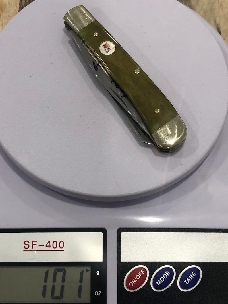 Op de afbeelding: Een gesloten zakmes met een groene handgreep en zilveren accenten, liggend op een weegschaal. Het mes heeft een klein embleem en een zichtbaar lemmet. De weegschaal geeft een gewicht van 286 gram aan.