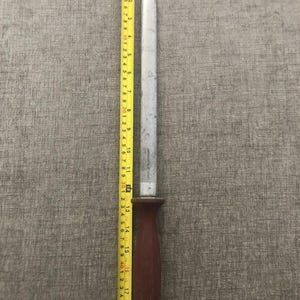 Pode incluir: Um afiador de facas de a&ccedil;o com um cabo castanho escuro, medindo aproximadamente 48 cm de comprimento, &eacute; mostrado ao lado de uma fita m&eacute;trica amarela. O afiador tem uma l&acirc;mina prateada e uma argola na extremidade do cabo.