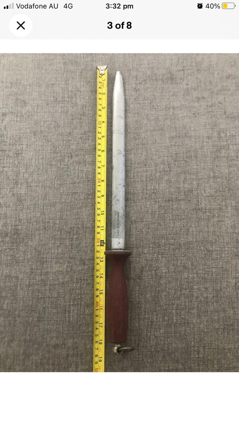 Pode incluir: Um afiador de facas de a&ccedil;o com um cabo castanho escuro, medindo aproximadamente 48 cm de comprimento, &eacute; mostrado ao lado de uma fita m&eacute;trica amarela. O afiador tem uma l&acirc;mina prateada e uma argola na extremidade do cabo.