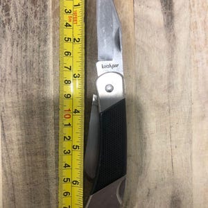 Puede incluir: Una navaja plegable plateada y negra con la marca "Kershaw" visible. El cuchillo est&aacute; junto a una cinta m&eacute;trica amarilla, que muestra una longitud de aproximadamente 18 cm.