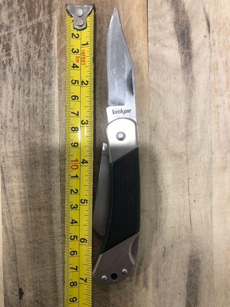 Puede incluir: Una navaja plegable plateada y negra con la marca "Kershaw" visible. El cuchillo est&aacute; junto a una cinta m&eacute;trica amarilla, que muestra una longitud de aproximadamente 18 cm.