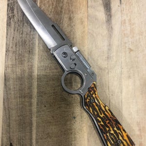 May include: A stainless steel folding knife with a long, sharp blade. The handle features a brown and yellow patterned design. The knife has a finger ring and a locking mechanism. The blade is approximately 10 cm long.