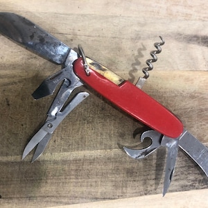 May include: A red-handled multi-tool, including a knife, scissors, corkscrew, and other implements. The metal parts show wear. This tool is designed for a variety of uses and tasks.