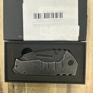 Könnte beinhalten: Ein dunkelgraues Klappmesser mit verwittertem Finish, strukturiertem Metallgriff und sichtbaren Schrauben, das in einer schwarzen, mit Schaumstoff ausgekleideten rechteckigen Schachtel liegt. Ein weißes Versandetikett mit Barcode klebt auf dem Deckel.