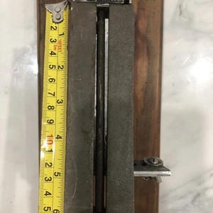 May include: A vintage metal tool with a wooden base, featuring a measuring tape showing approximately 7 inches. The tool has a rectangular shape with a central channel and a screw mechanism. The metal has a worn, aged appearance.