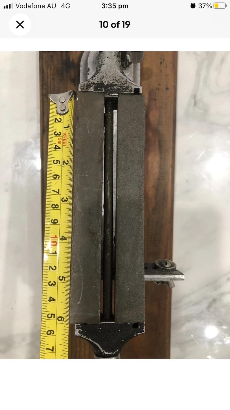 May include: A vintage metal tool with a wooden base, featuring a measuring tape showing approximately 7 inches. The tool has a rectangular shape with a central channel and a screw mechanism. The metal has a worn, aged appearance.