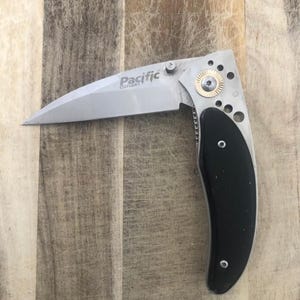 May include: A silver folding knife with a curved black handle. The blade is open, displaying the text "Pacific Cutlery". The knife has a gold-coloured accent near the hinge and several small holes in the metal.