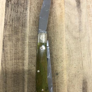 Op de afbeelding: Een zakmes met een roestvrijstalen lemmet en een groene handgreep. De handgreep heeft een messing bolster en drie klinknagels. Het mes is open, waardoor het lemmet zichtbaar is, tegen een houten achtergrond.