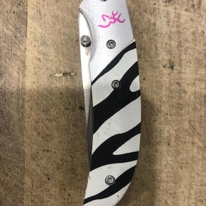 K&ouml;nnte beinhalten: Ein Klappmesser mit silberfarbenem Metallgriff und einem Zebra-Streifen-Design in Schwarz und Wei&szlig;. Das Messer hat ein rosa Logo auf dem Griff und ist geschlossen. Das Messer liegt auf einer Holzoberfl&auml;che.