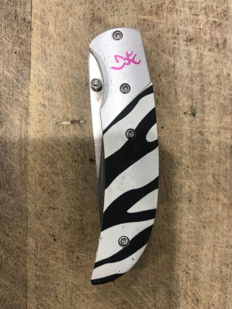 K&ouml;nnte beinhalten: Ein Klappmesser mit silberfarbenem Metallgriff und einem Zebra-Streifen-Design in Schwarz und Wei&szlig;. Das Messer hat ein rosa Logo auf dem Griff und ist geschlossen. Das Messer liegt auf einer Holzoberfl&auml;che.