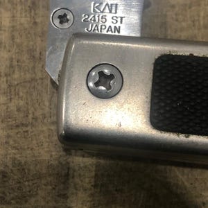 May include: Close-up of a silver metal knife with the text "KAI 2415 ST JAPAN" etched on the blade. The handle is also silver with a black textured grip. Two screws are visible.