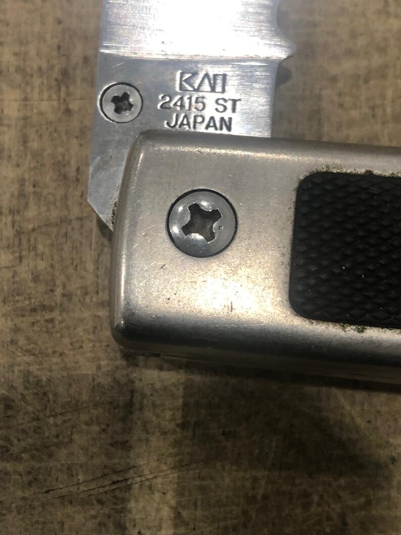 May include: Close-up of a silver metal knife with the text "KAI 2415 ST JAPAN" etched on the blade. The handle is also silver with a black textured grip. Two screws are visible.