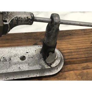 May include: Close-up of a vintage metal tool with a silver finish, mounted on a wooden surface. The tool features a curved base, a central hole, and a rotating knob. A metal rod extends across the top, connecting to a cylindrical component.