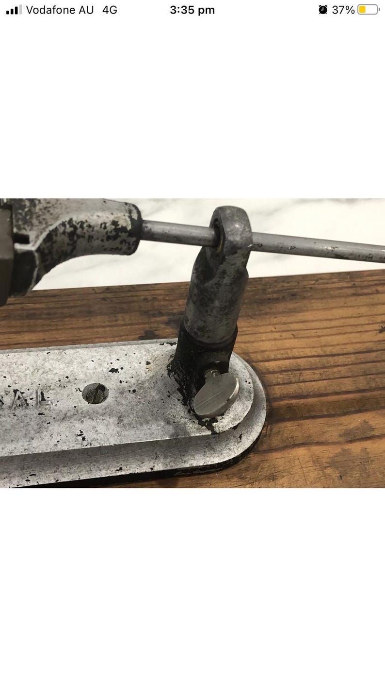 May include: Close-up of a vintage metal tool with a silver finish, mounted on a wooden surface. The tool features a curved base, a central hole, and a rotating knob. A metal rod extends across the top, connecting to a cylindrical component.
