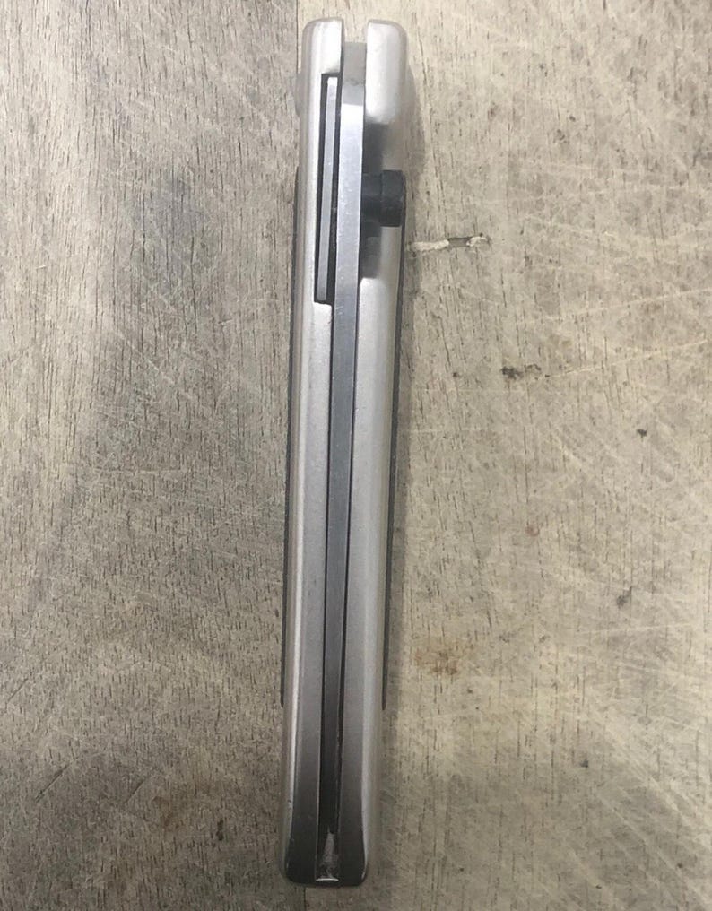 Pode incluir: Uma faca dobr&aacute;vel prateada fechada com um parafuso preto. A faca tem uma forma retangular e um acabamento met&aacute;lico liso. A l&acirc;mina n&atilde;o &eacute; vis&iacute;vel nesta imagem.