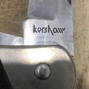Puede incluir: Primer plano de una navaja plegable parcialmente abierta con una hoja plateada. La palabra "Kershaw" est&aacute; grabada en la hoja. El mango es una combinaci&oacute;n de metal plateado y material negro. El cuchillo est&aacute; sobre una superficie de madera.