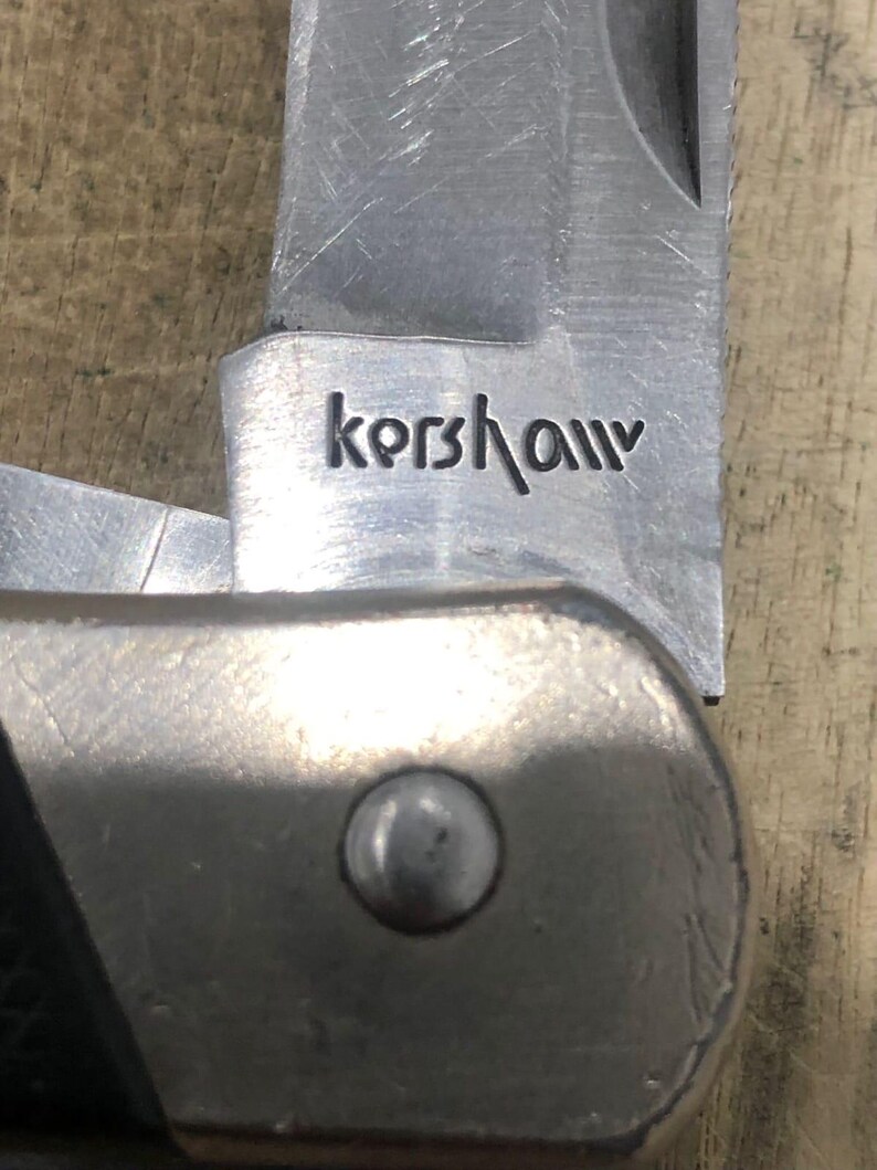 Puede incluir: Primer plano de una navaja plegable parcialmente abierta con una hoja plateada. La palabra "Kershaw" est&aacute; grabada en la hoja. El mango es una combinaci&oacute;n de metal plateado y material negro. El cuchillo est&aacute; sobre una superficie de madera.