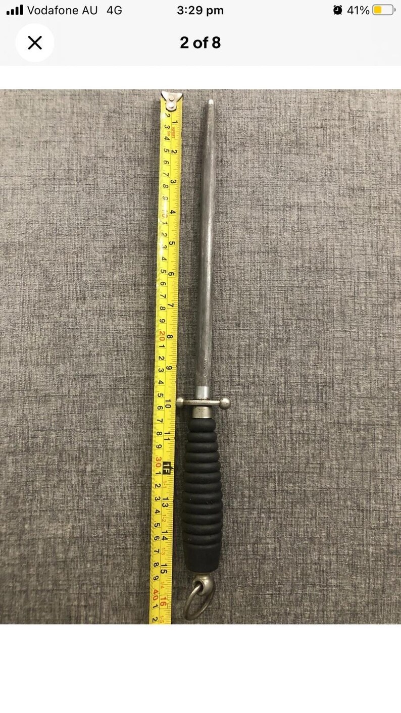 May include: A steel knife sharpener with a black handle and a ring at the end. The sharpener is approximately 16 inches long. A yellow measuring tape is placed next to the sharpener.