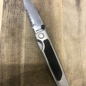 May include: A silver folding knife with a partially serrated blade. The handle has a black rubber grip and the brand name "Kershaw". The knife is on a wooden surface.