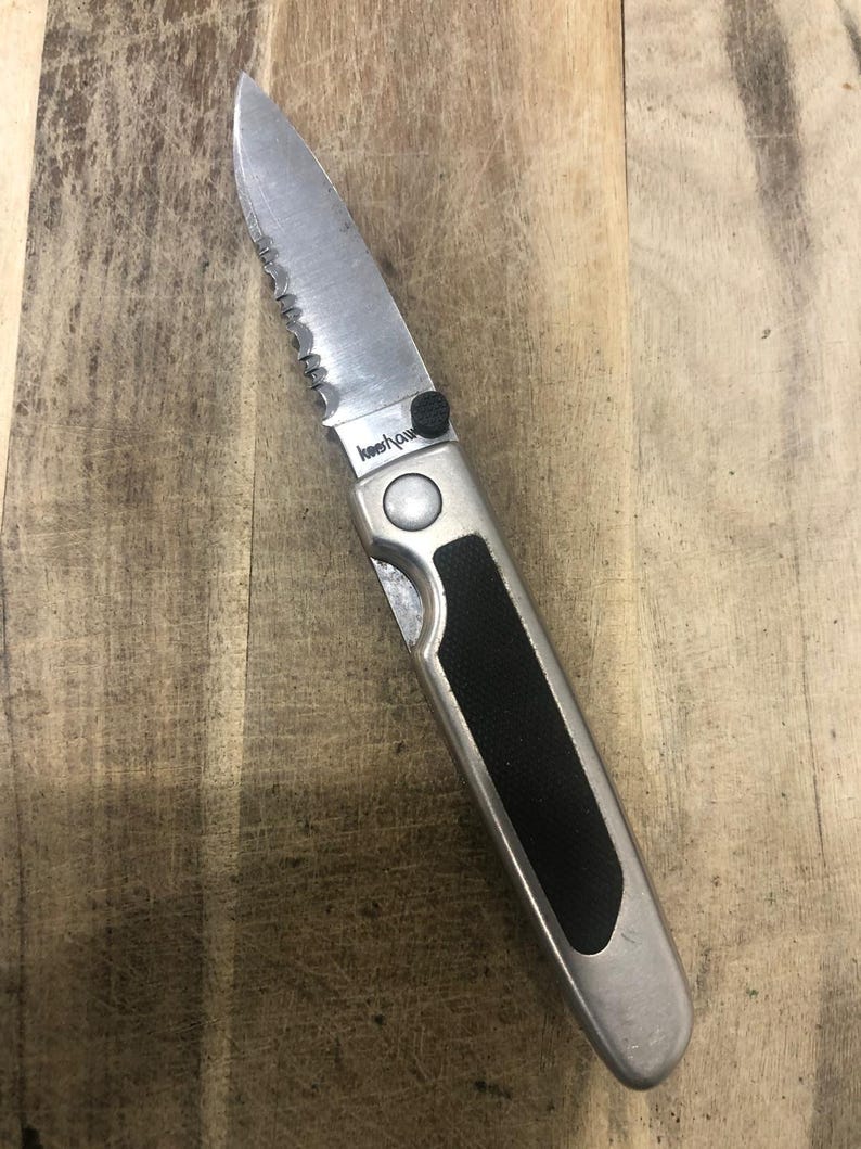 May include: A silver folding knife with a partially serrated blade. The handle has a black rubber grip and the brand name "Kershaw". The knife is on a wooden surface.
