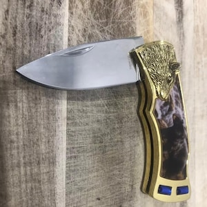 Puede incluir: Una navaja plegable de acero inoxidable con una hoja pulida. El mango presenta un diseño de cabeza de oso dorado y una inserción estampada con detalles azules. El cuchillo está sobre un fondo de madera.