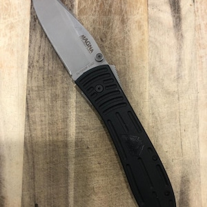 Peut inclure: Un couteau pliant noir et argenté avec le texte "MAGNA" et "OUTDOOR EDGE" sur la lame. Le couteau a un manche noir texturé et une lame argentée. Le couteau est posé sur une surface en bois.