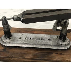 May include: A vintage metal tool with the text "UNIVERSAL" stamped on the base. The tool has a dark, rectangular top section and is supported by two upright metal supports. The base is silver-colored and rests on a wooden surface.