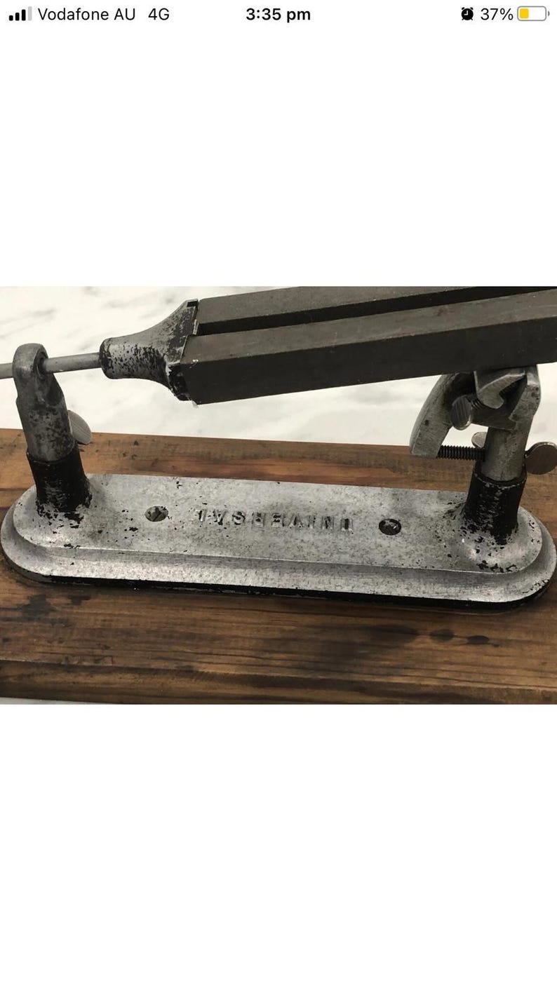 May include: A vintage metal tool with the text "UNIVERSAL" stamped on the base. The tool has a dark, rectangular top section and is supported by two upright metal supports. The base is silver-colored and rests on a wooden surface.