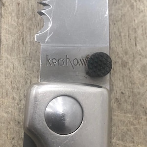 Pode incluir: Close-up de uma faca dobr&aacute;vel prateada com uma l&acirc;mina parcialmente serrilhada. A palavra "kershaw" est&aacute; gravada na l&acirc;mina. A pega &eacute; prateada com uma pega texturizada preta. Uma ferramenta pr&aacute;tica.