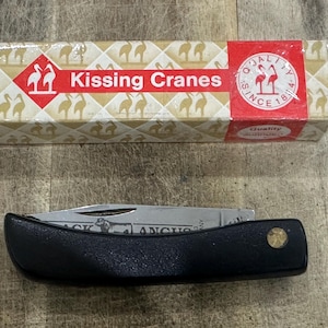 May include: A folding pocket knife with a black handle and a metallic blade, positioned below its rectangular cardboard packaging. The box features a repeating crane pattern, a red banner with the text Kissing Cranes, and a circular logo.