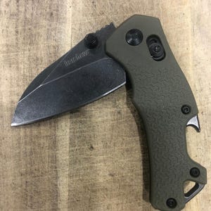 May include: An open, olive-green folding knife with a black blade. The blade is partially exposed, revealing the word "Kershaw." The handle has a textured pattern and includes a bottle opener and a small hole. The knife is set against a wooden surface.