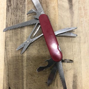 May include: A red multi-tool with various stainless steel implements extended, including a knife, saw, scissors, and bottle opener. The tool is open on a wooden surface, showcasing its versatility.