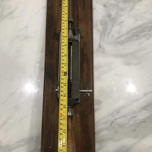 May include: A vintage metal and wood tool, possibly for sharpening, mounted on a dark wooden board. A yellow measuring tape shows the tool is approximately 20 inches long. The tool has a metal blade and handle.