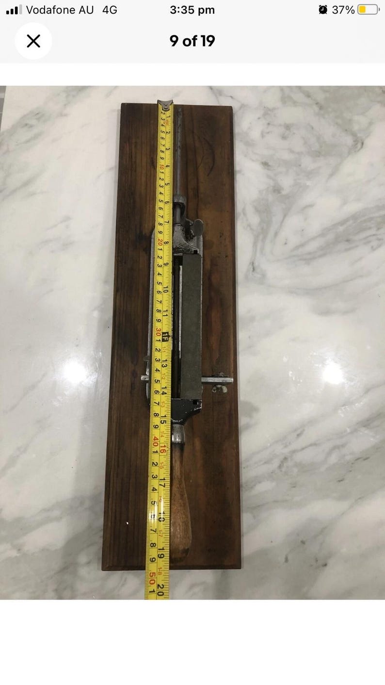 May include: A vintage metal and wood tool, possibly for sharpening, mounted on a dark wooden board. A yellow measuring tape shows the tool is approximately 20 inches long. The tool has a metal blade and handle.