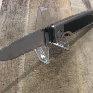 May include: A silver and black folding knife with a serrated blade is displayed on a clear plastic stand. The knife has a textured black grip and a silver blade. The handle has a silver button. The knife is on a wooden surface.