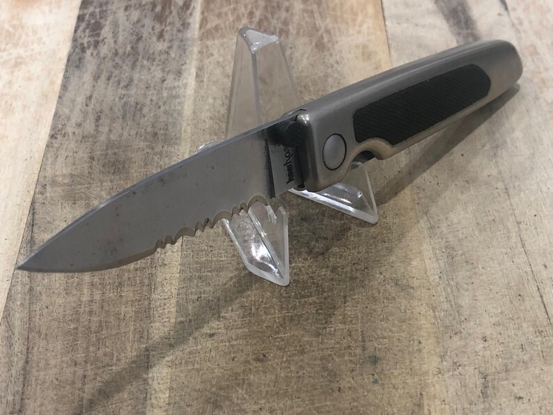 May include: A silver and black folding knife with a serrated blade is displayed on a clear plastic stand. The knife has a textured black grip and a silver blade. The handle has a silver button. The knife is on a wooden surface.