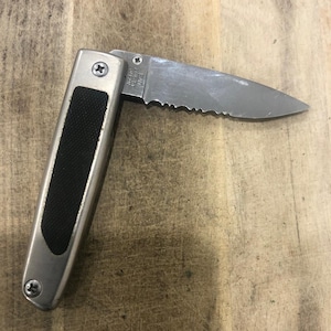 May include: A silver folding knife with a partially serrated blade. The handle is silver with a black rubber grip. The blade is open, showing the serrated edge. The knife is on a wooden surface.