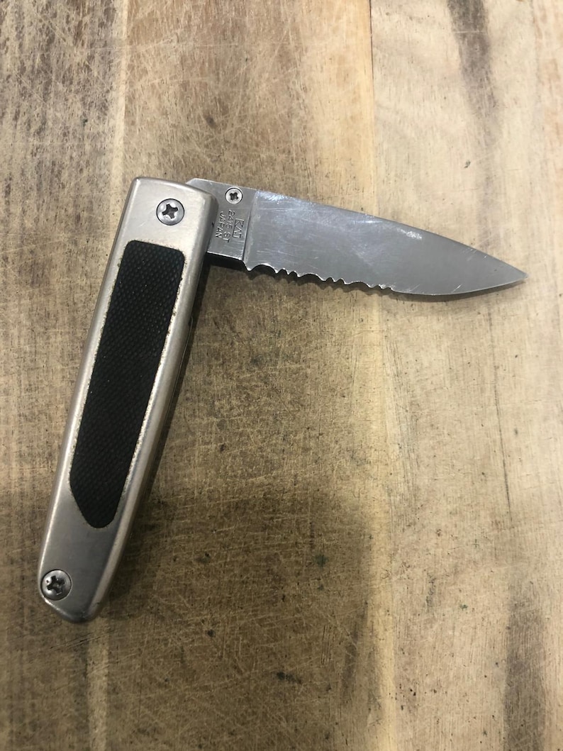 May include: A silver folding knife with a partially serrated blade. The handle is silver with a black rubber grip. The blade is open, showing the serrated edge. The knife is on a wooden surface.