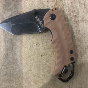 May include: A tan and black folding knife with the brand name "Kershaw" printed on the blade. The blade is dark grey and partially open. The handle is tan with a textured surface and a black clip.