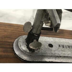 May include: Close-up of a vintage metal tool with a silver finish. The tool features adjustable knobs and a curved base, mounted on a wooden surface. The design suggests a mechanical or industrial purpose.