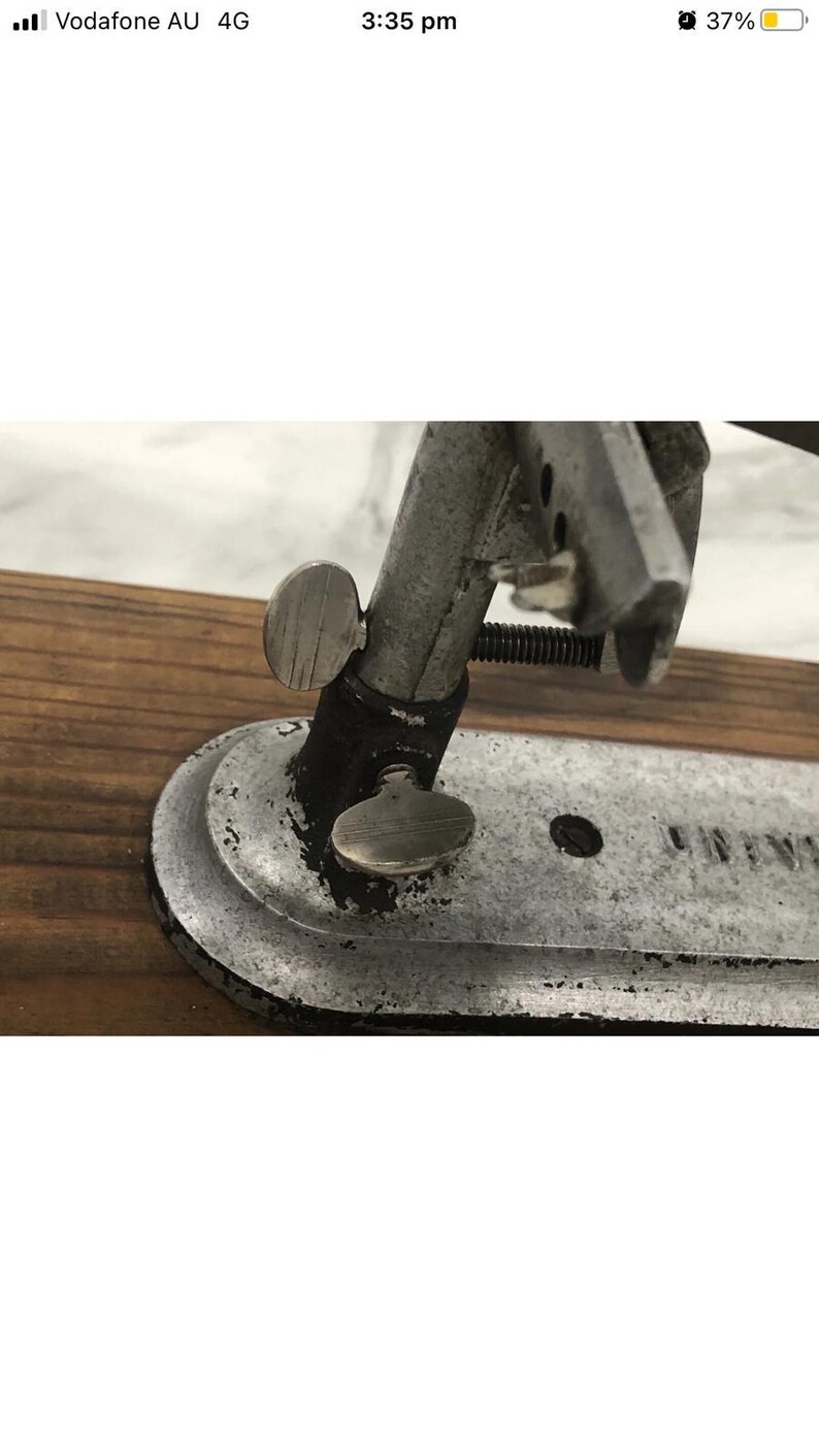 May include: Close-up of a vintage metal tool with a silver finish. The tool features adjustable knobs and a curved base, mounted on a wooden surface. The design suggests a mechanical or industrial purpose.