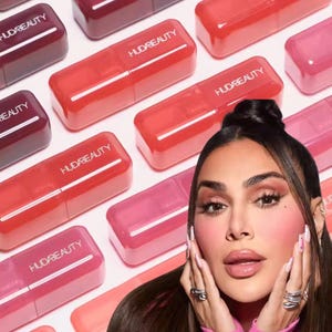 May include: An assortment of Huda Beauty lip products in various shades of red and pink. The rectangular tubes are arranged on a white surface. The brand name is printed on each tube. A woman is in the foreground, with her hands near her face.