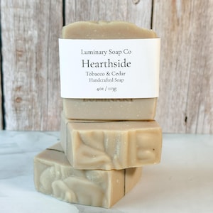 May include: A stack of three rectangular bars of light tan soap. The top bar features a white paper label with black text reading Luminary Soap Co, Hearthside, Tobacco & Cedar, Handcrafted Soap, 4oz / 113g. The soap has a smooth, matte texture.